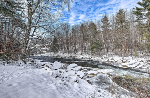 A snowy landscape with a river flowing through trees, under a blue sky with scattered clouds.