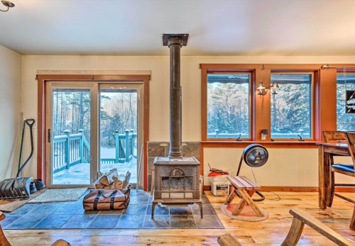 Cozy interior with a wood stove, wooden floors, and large windows overlooking a snowy landscape.