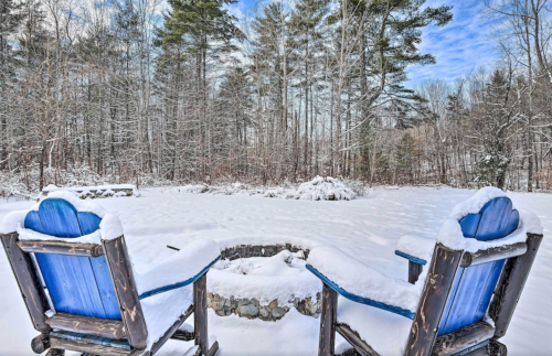 Two blue chairs covered in snow, facing a stone fire pit in a snowy forest landscape.