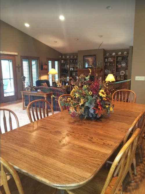 A wooden dining table with a floral centerpiece in a cozy living room with bookshelves and soft lighting.