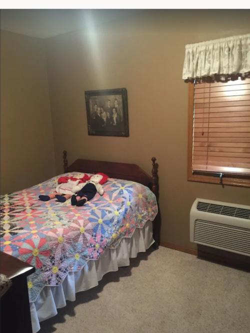 Cozy bedroom with a quilted bedspread, a window with blinds, and a framed picture on the wall.