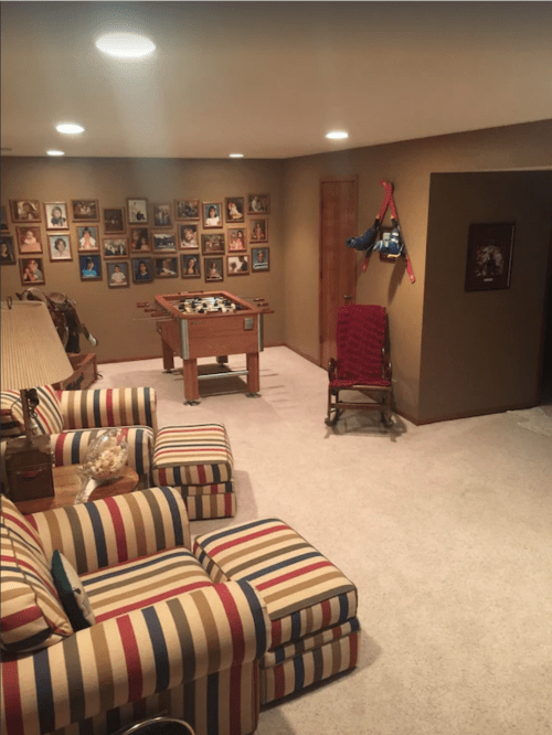 Cozy basement with striped sofas, a foosball table, and a wall of framed photos. Soft lighting and carpeted floor.