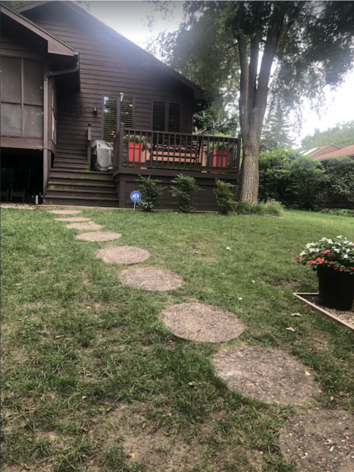 A pathway of stone steps leads to a brown house with a porch, surrounded by greenery and potted plants.