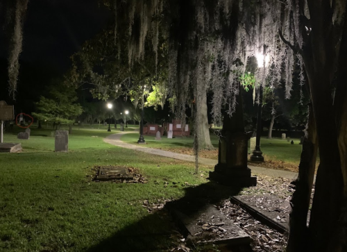 A dimly lit cemetery at night, featuring gravestones, trees with Spanish moss, and a winding path illuminated by lamps.
