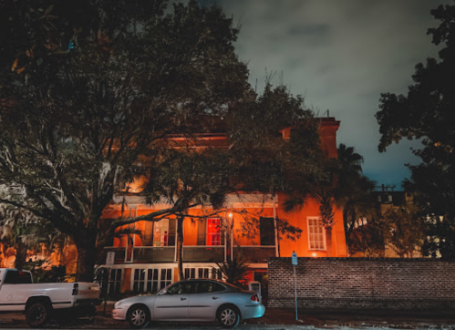 A historic building illuminated at night, surrounded by trees and parked cars under a cloudy sky.