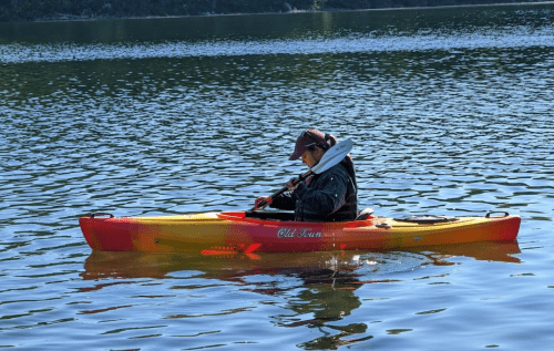 A person in a kayak paddles on a calm lake, surrounded by water and trees.
