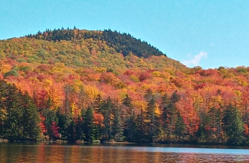 Vibrant autumn foliage reflects on a calm lake, with a forested hill in the background under a clear blue sky.