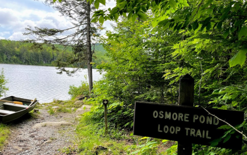 A serene view of Osmore Pond Loop Trail with a wooden sign and a small boat by the water's edge, surrounded by greenery.