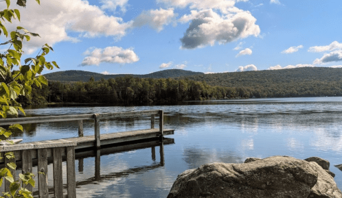A serene lake scene with a wooden dock, surrounded by trees and mountains under a blue sky with fluffy clouds.