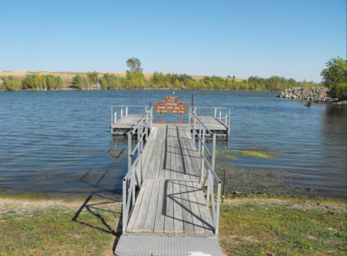 A wooden dock extends into a calm lake, surrounded by greenery and clear blue skies.