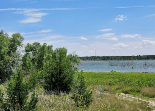 A serene landscape featuring a calm lake, green vegetation, and a clear blue sky with scattered clouds.