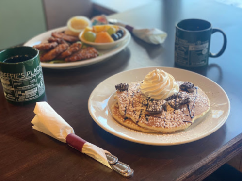 A plate of pancakes topped with whipped cream and chocolate, with a side of fruit and chicken wings in the background.