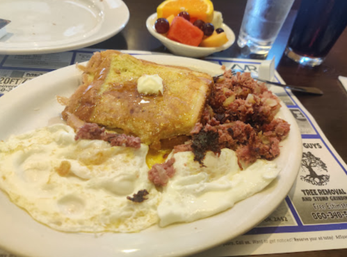 A plate of breakfast featuring French toast with butter, scrambled eggs, and a side of hash browns. Fresh fruit on the side.