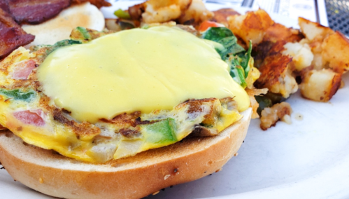 A close-up of a sandwich with a cheesy egg patty, spinach, and tomatoes, served with crispy hash browns on the side.