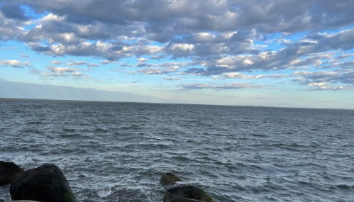 A serene seascape with gentle waves, rocky shoreline, and a sky filled with clouds.