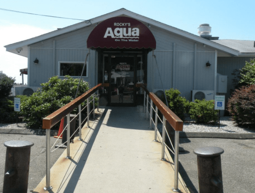 Entrance to Rocky's Aqua on the Water, featuring a ramp and a sign above the door. Lush greenery surrounds the building.