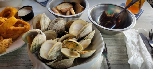 A bowl of clams with bread and onion rings on a table, accompanied by dipping sauces and a drink.