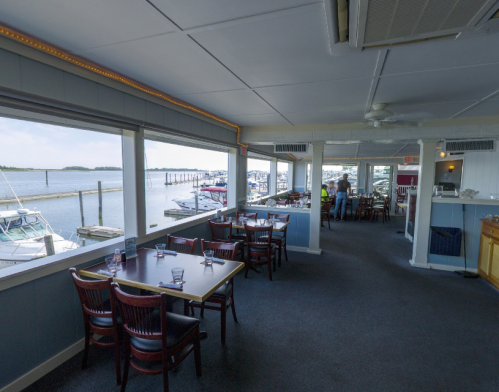 Interior of a waterfront restaurant with tables, large windows, and a view of boats in the marina.