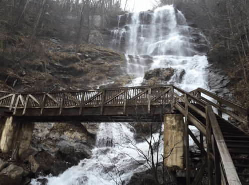 A wooden bridge crosses over a waterfall, surrounded by rocky terrain and bare trees in a misty landscape.