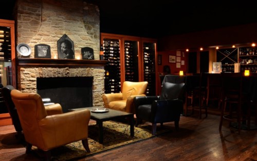 Cozy lounge with leather chairs, a stone fireplace, and a wine cellar in the background, softly lit for ambiance.