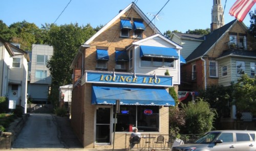 A brick building with blue awnings, featuring a sign that reads "Lounge 110," surrounded by trees and other houses.