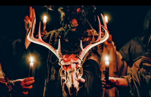 A dark scene featuring a deer skull with antlers, surrounded by figures holding candles in a ritualistic setting.