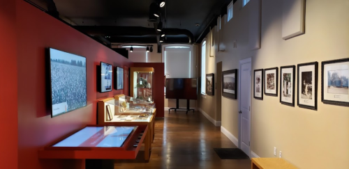Interior of a museum exhibit featuring displays, photographs, and interactive screens along a red wall.