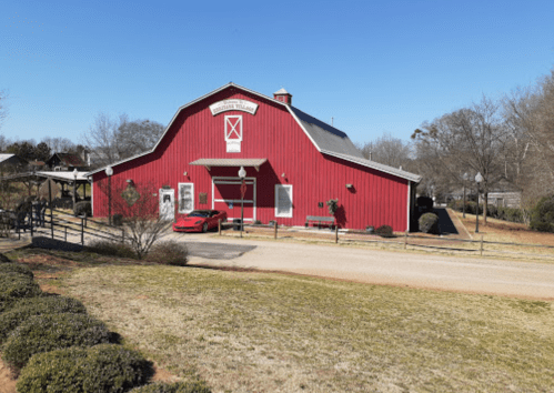 A red barn-style building with a clear blue sky, surrounded by grass and trees. A red sports car is parked in front.