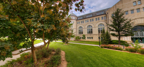 A landscaped area with trees and green grass beside a large stone building under a cloudy sky.