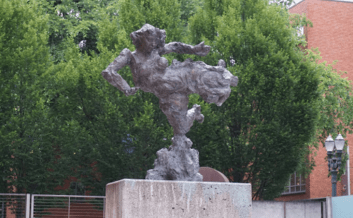 A bronze sculpture of a dynamic figure in motion, set against a backdrop of green trees and a brick building.