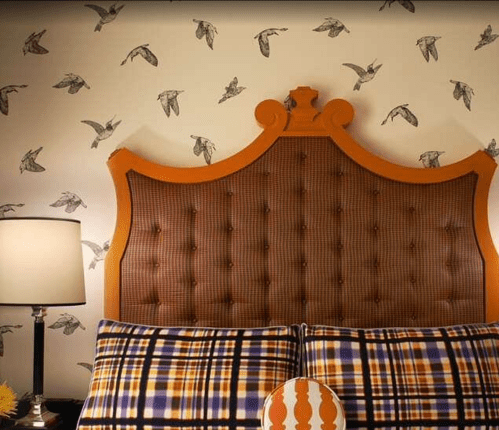 A stylish bedroom featuring a tufted orange headboard, plaid pillows, and bird-patterned wallpaper.