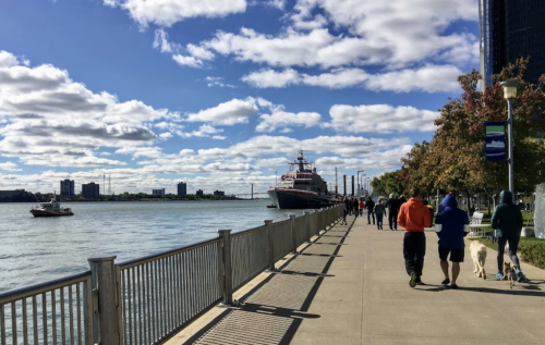 People walking along a waterfront path with a boat and a large ship in the background under a partly cloudy sky.
