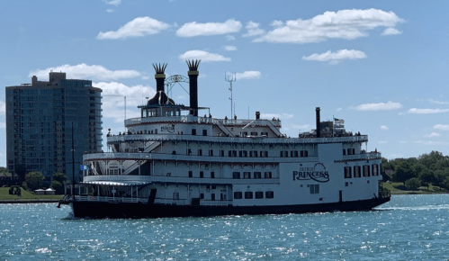 A large riverboat named "Princess" sails on a blue waterway, with a city skyline in the background under a clear sky.