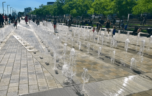 Children and adults play in a splash pad with water jets, surrounded by trees and park benches on a sunny day.