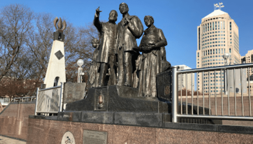 A bronze statue of three historical figures stands in a park, with modern buildings in the background.
