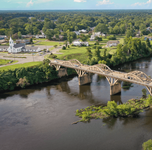 Aerial view of a bridge over a river, surrounded by greenery and a small town with a church in the background.