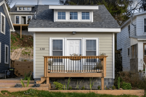 A small, modern house with a front porch, white door, and gray roof, surrounded by greenery.