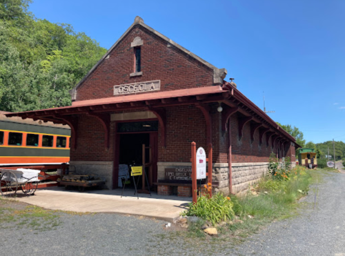 Historic brick train station in Osceola, surrounded by greenery and vintage train cars. Clear blue sky above.