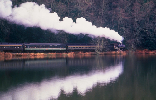 A steam train travels along a river, with smoke billowing and trees reflected in the calm water.