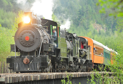 A vintage steam train with a black locomotive and orange passenger car travels over a wooden bridge in a lush green landscape.