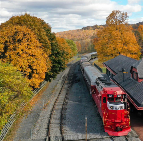 A red train passes through a scenic landscape with autumn trees and a vintage station under a blue sky.