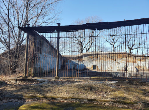 A rusted fence surrounds a dilapidated building, with bare trees and a clear blue sky in the background.