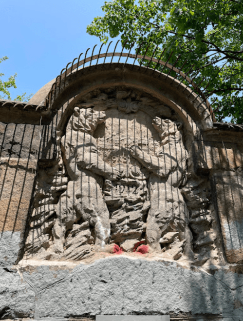 Relief sculpture on a stone wall depicting two figures, surrounded by greenery and a clear blue sky.