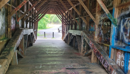 Interior of a covered bridge with colorful graffiti on wooden beams and walls, leading to an open area outside.