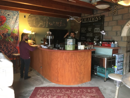 A cozy bar with a wooden counter, a bartender, and a customer, surrounded by colorful decor and a menu board.