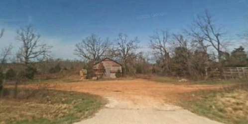 A dirt road leads to a rustic barn surrounded by bare trees and open land under a clear blue sky.