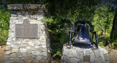 A stone monument with a plaque beside a large bell, surrounded by greenery.