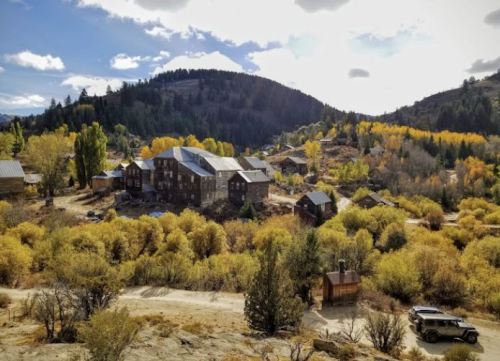 A scenic view of a rustic town surrounded by autumn foliage and mountains under a partly cloudy sky.