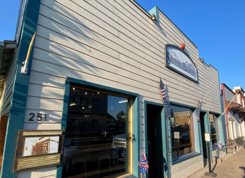 A storefront with a sign reading "Sisters Bakery," featuring patriotic decorations and a clear blue sky.
