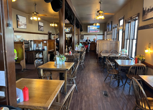 A cozy restaurant interior with wooden tables, chairs, and decorative flowers, featuring a bar area in the background.